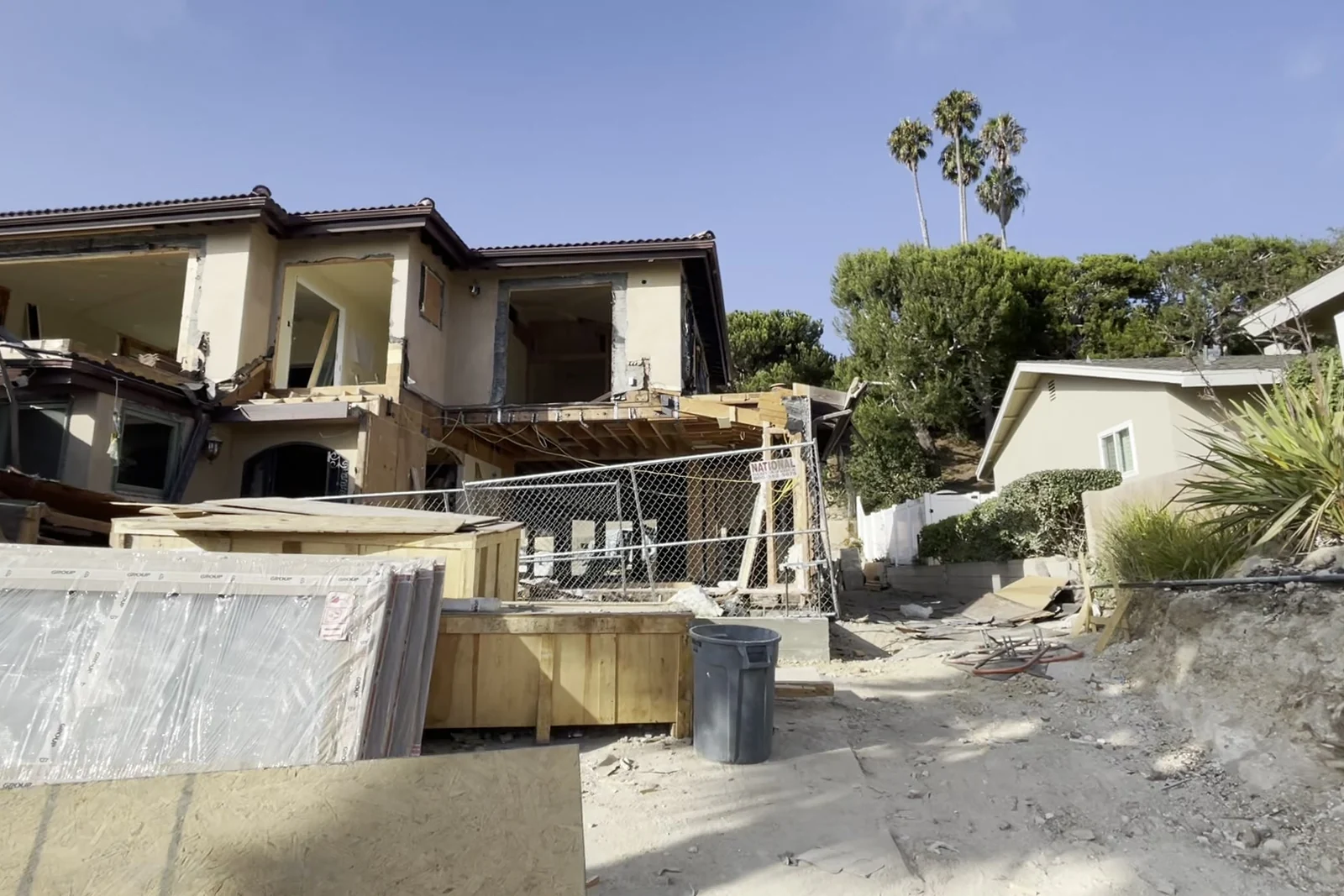 The house mid-works from outside on its terraced lot, roof opened and walls stripped, with the neighbouring rooflines and the hillside behind