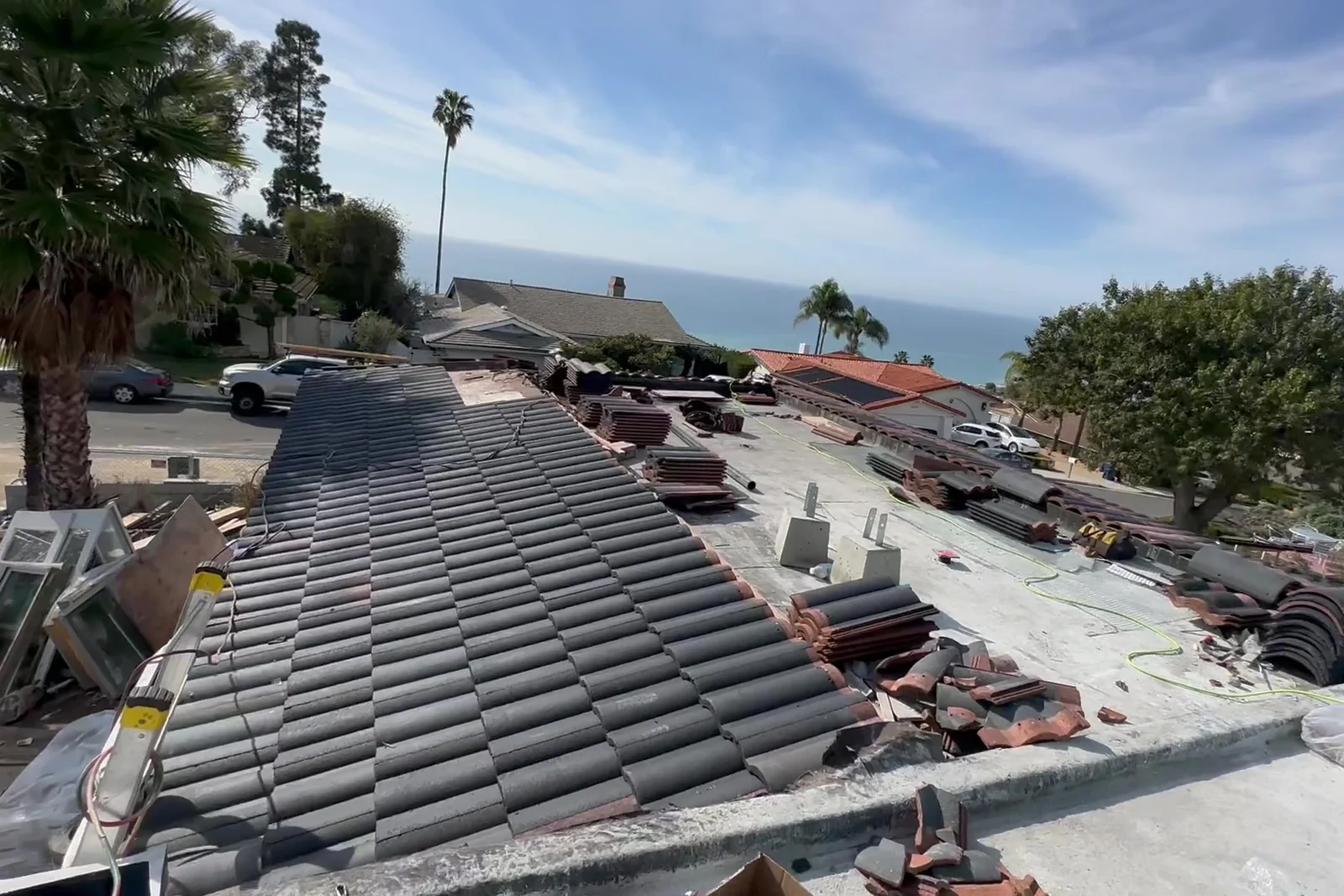 Original terracotta barrel tiles stacked in rows across the open roof deck as they come off, with palm tops and the ocean beyond