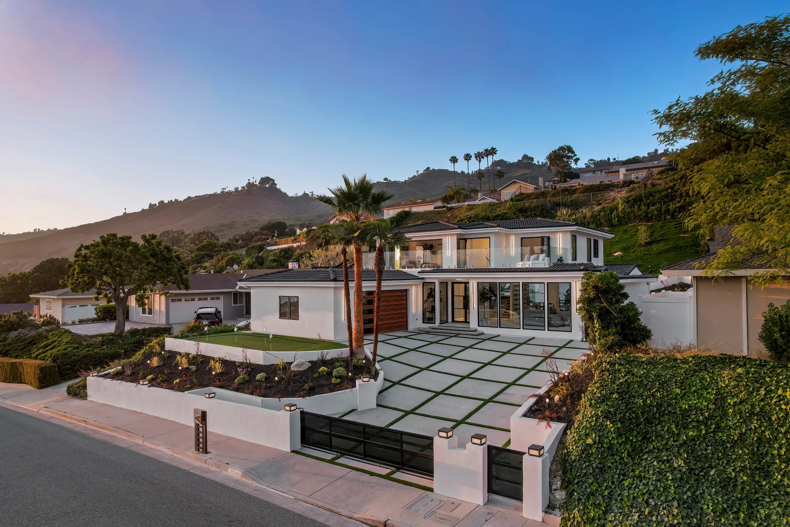 The same house at dusk in white stucco with a dark flat-profile tile roof, black-framed glass across the ground floor, a walnut slatted garage door, a glass-guarded upper balcony and a wide paver-and-turf motorcourt behind a low white wall