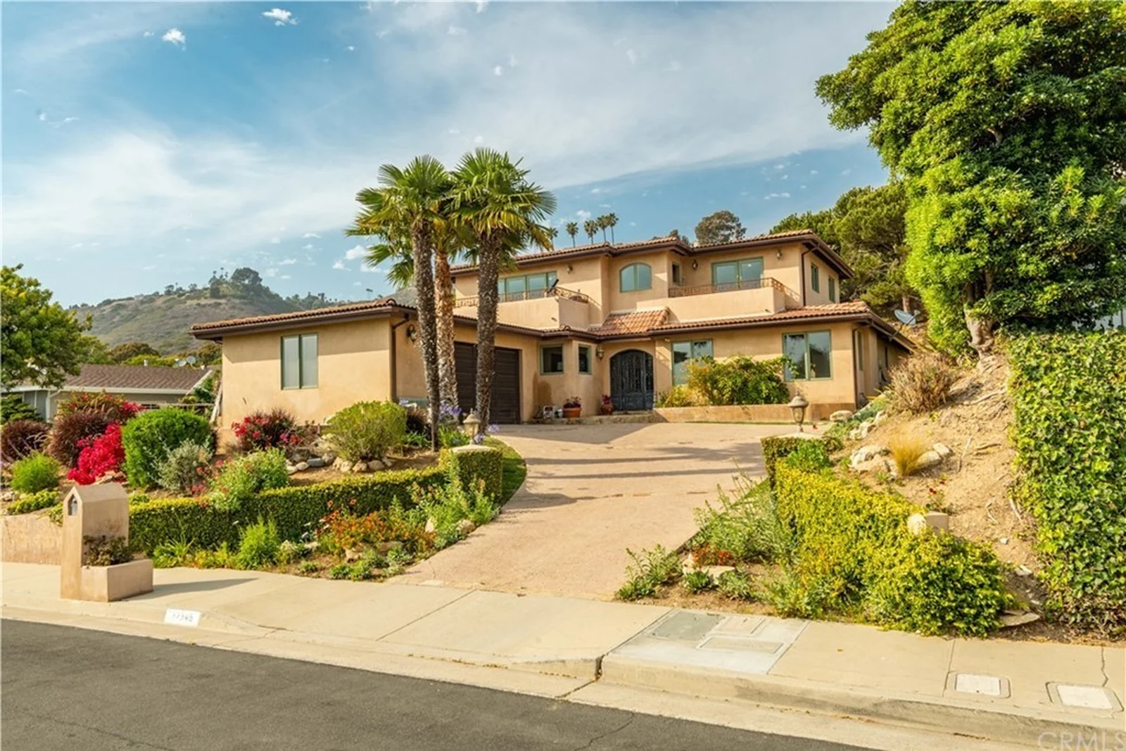 The original two-storey Mediterranean house in apricot stucco with a terracotta S-tile roof, green window frames, an arched iron entry door and a plain sloping driveway between deep mixed planting