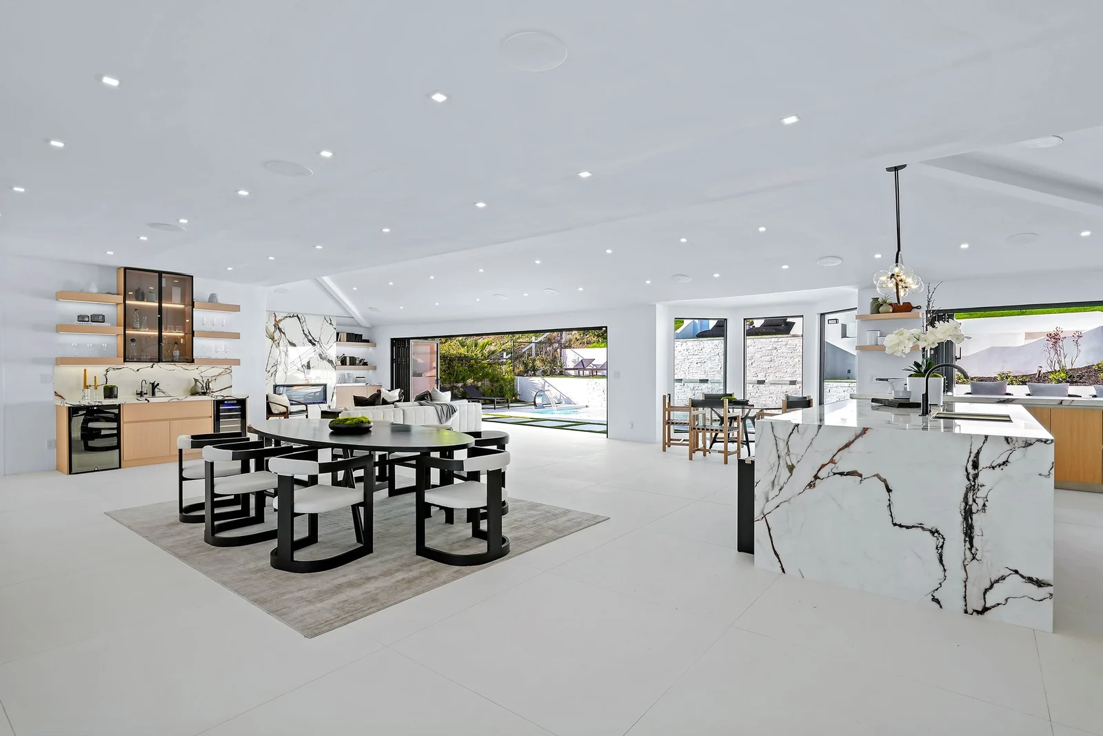 An open kitchen and dining space in white with marble slab surfaces, a round black table, a sculptural linear pendant, floating oak shelving and a clear sightline the length of the floor