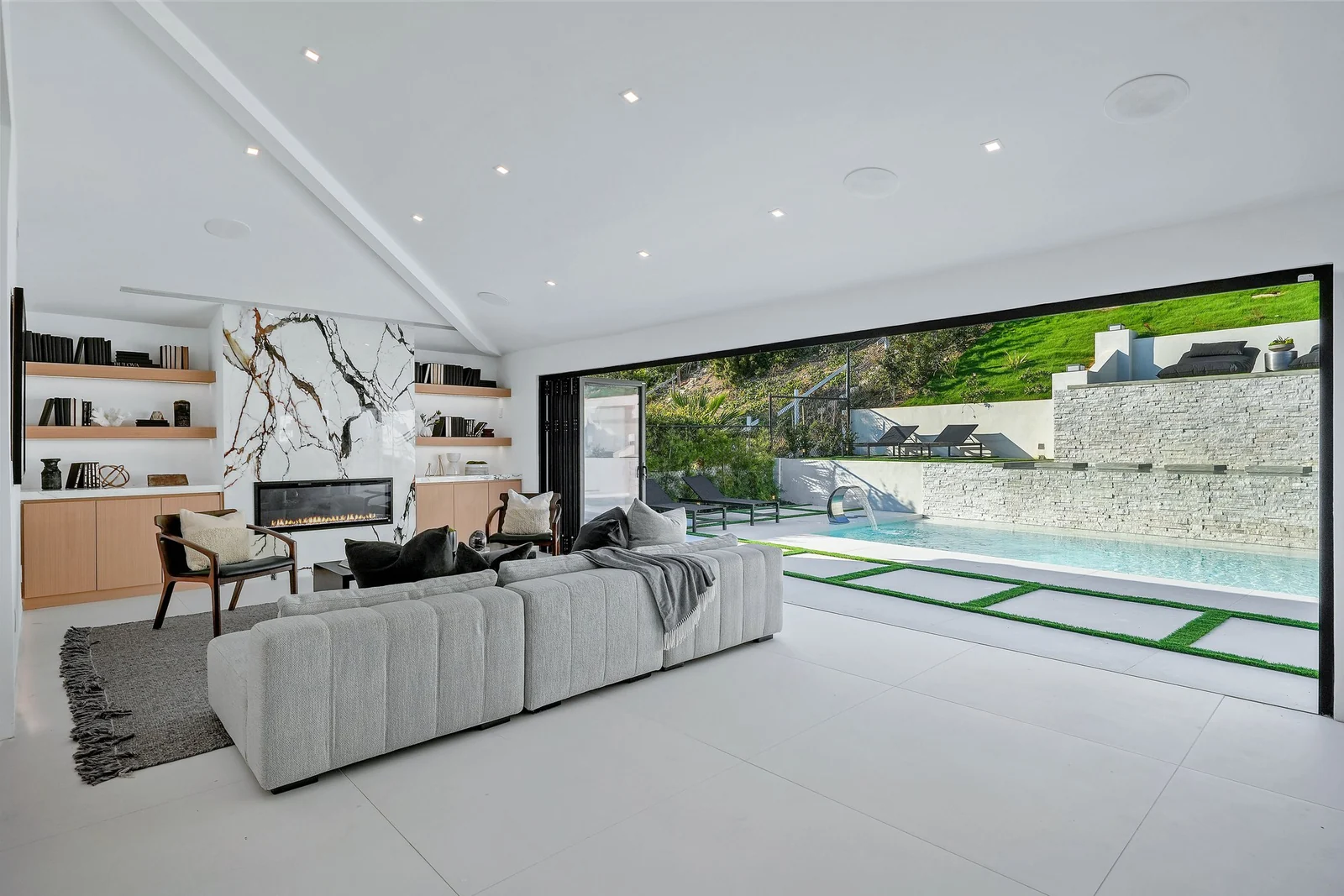 The same vaulted room now white with an exposed painted beam, a bookmatched marble fireplace panel, pale oak shelving and a black-framed glass wall folded fully open onto the pool terrace and green hillside
