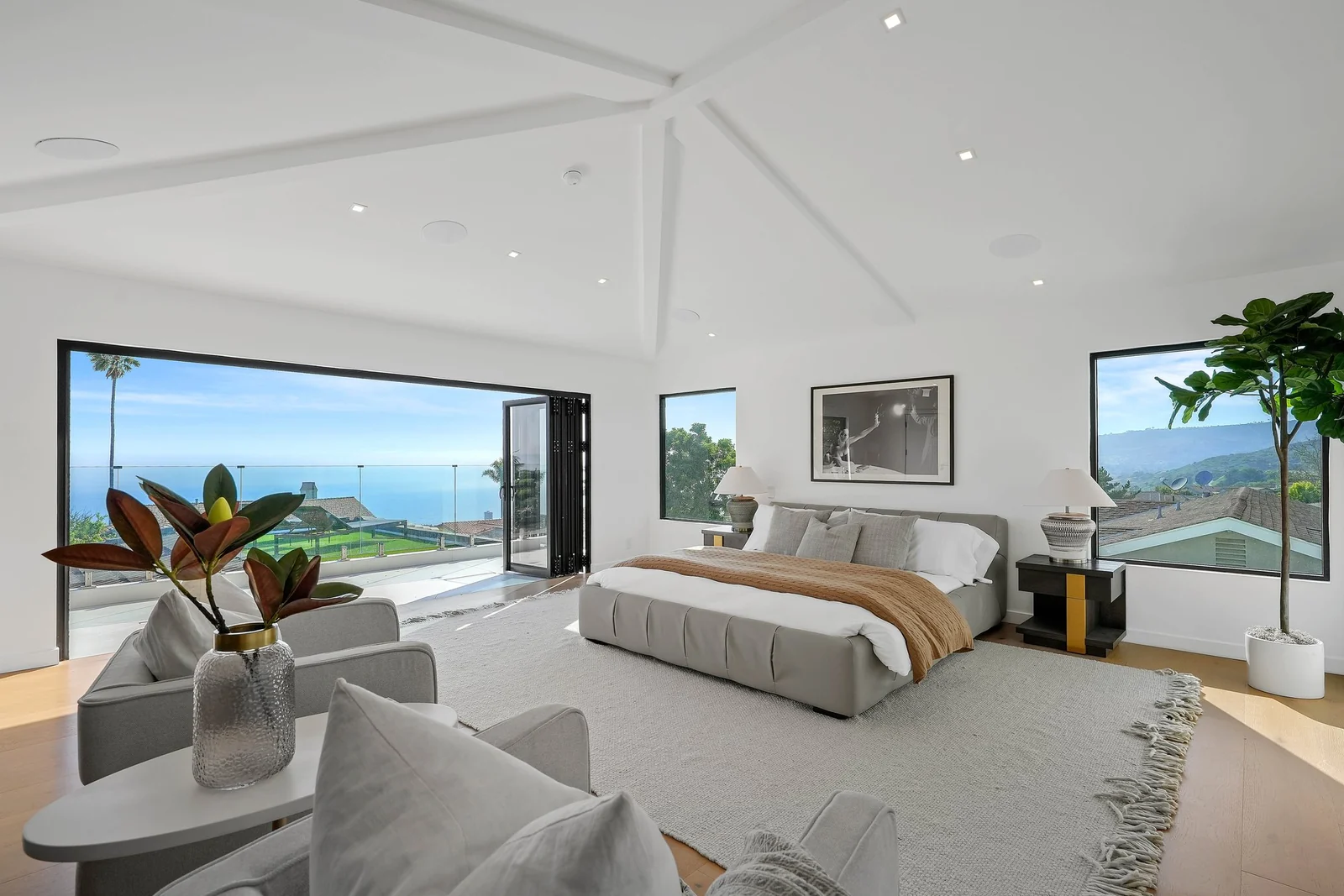 The primary suite with a white oak floor, a white vaulted ceiling with exposed painted rafters, a corner window onto the hillside and a full glass wall folded open to a balcony with a frameless glass guard and the ocean horizon beyond
