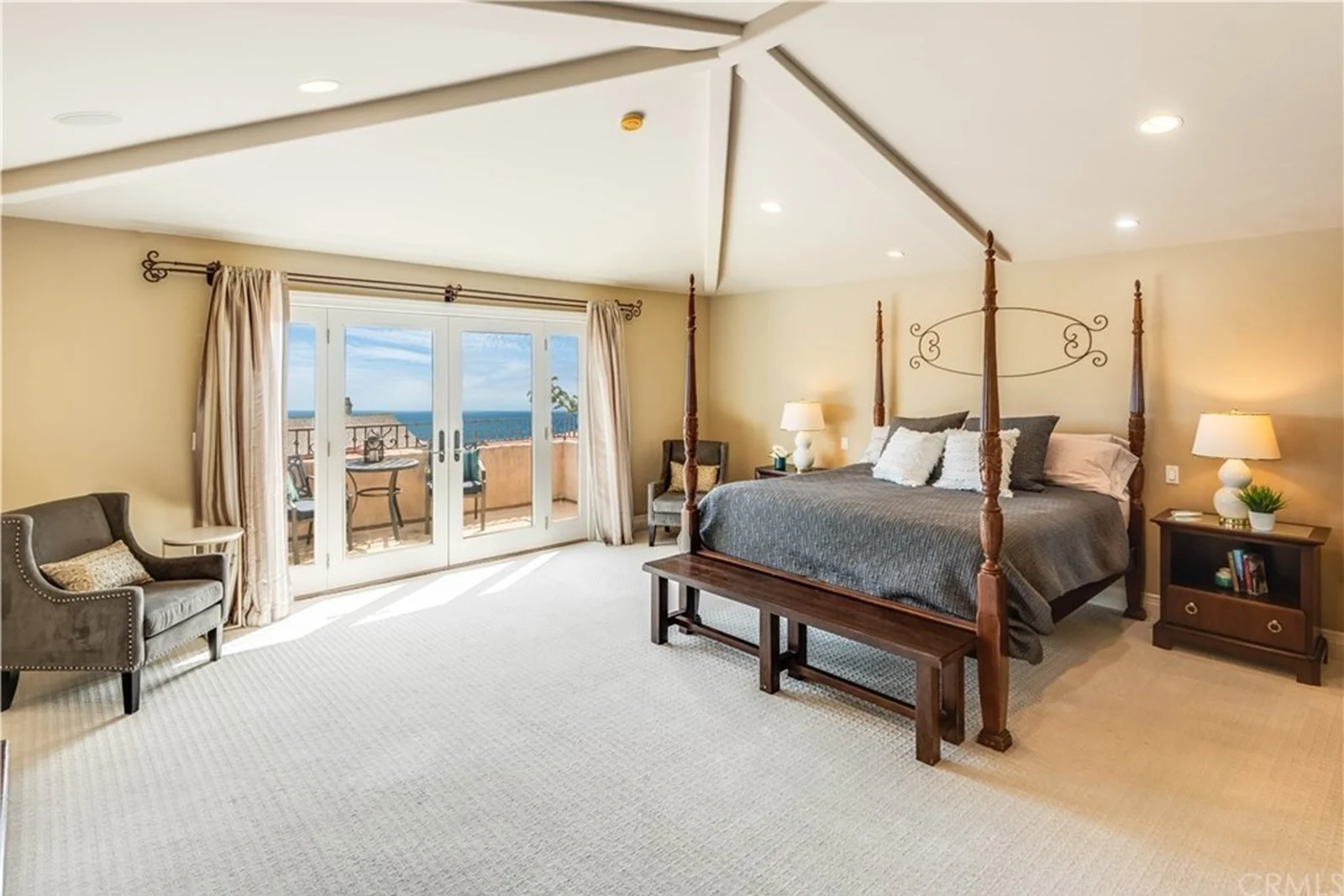 A primary bedroom with a dark carved four-poster bed, a wall-mounted television, carpet and a pair of French doors onto a small balcony