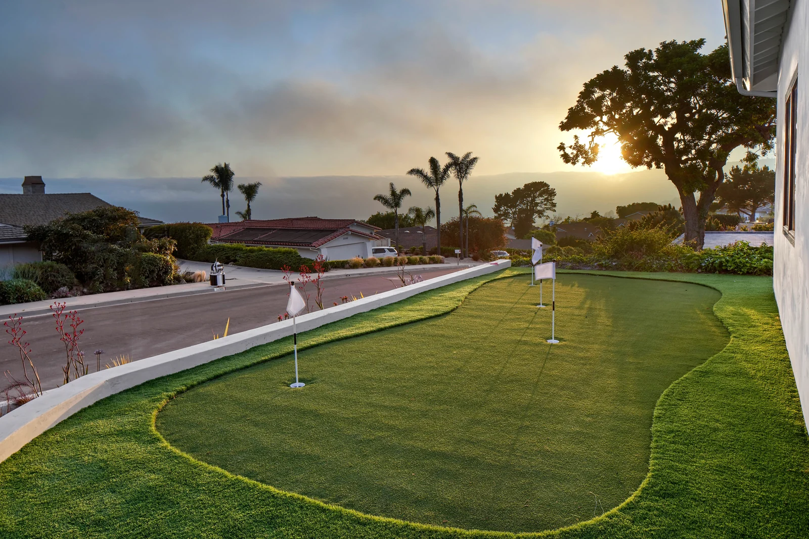 The same strip rebuilt as a curved putting green with three flags, edged by a low white wall and drought planting, looking west into the sunset over the coastline