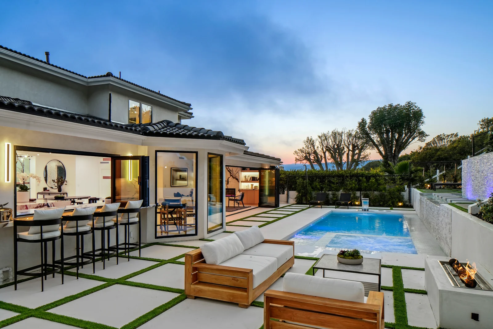 The same elevation at dusk with the glass folded fully open, a bar counter and stools under the kitchen window, teak lounge furniture, a linear gas fire, the pool lit blue and the sunset behind the trees
