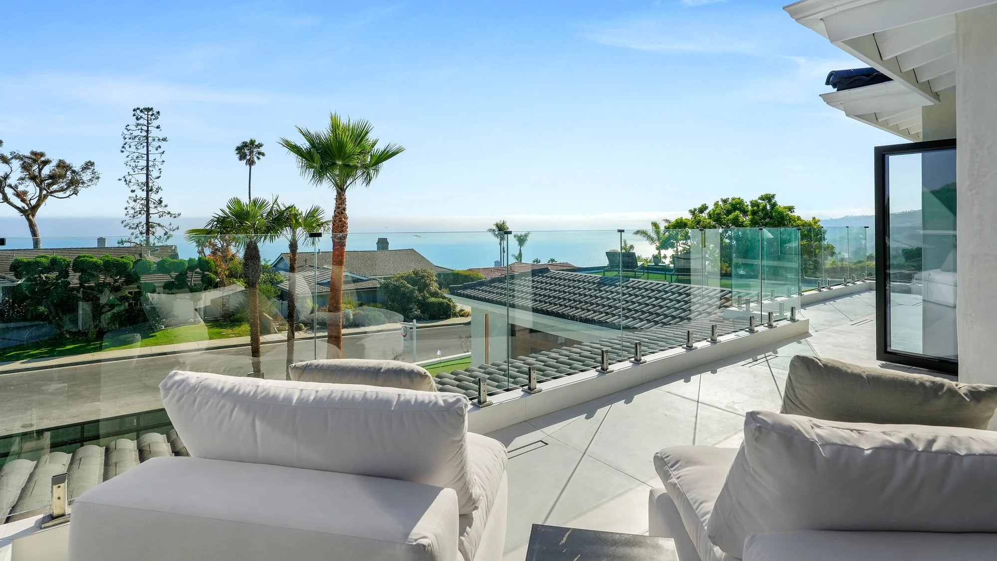Upper terrace with a frameless glass guard and white lounge chairs looking over neighbouring tile rooftops and palms to the open ocean horizon