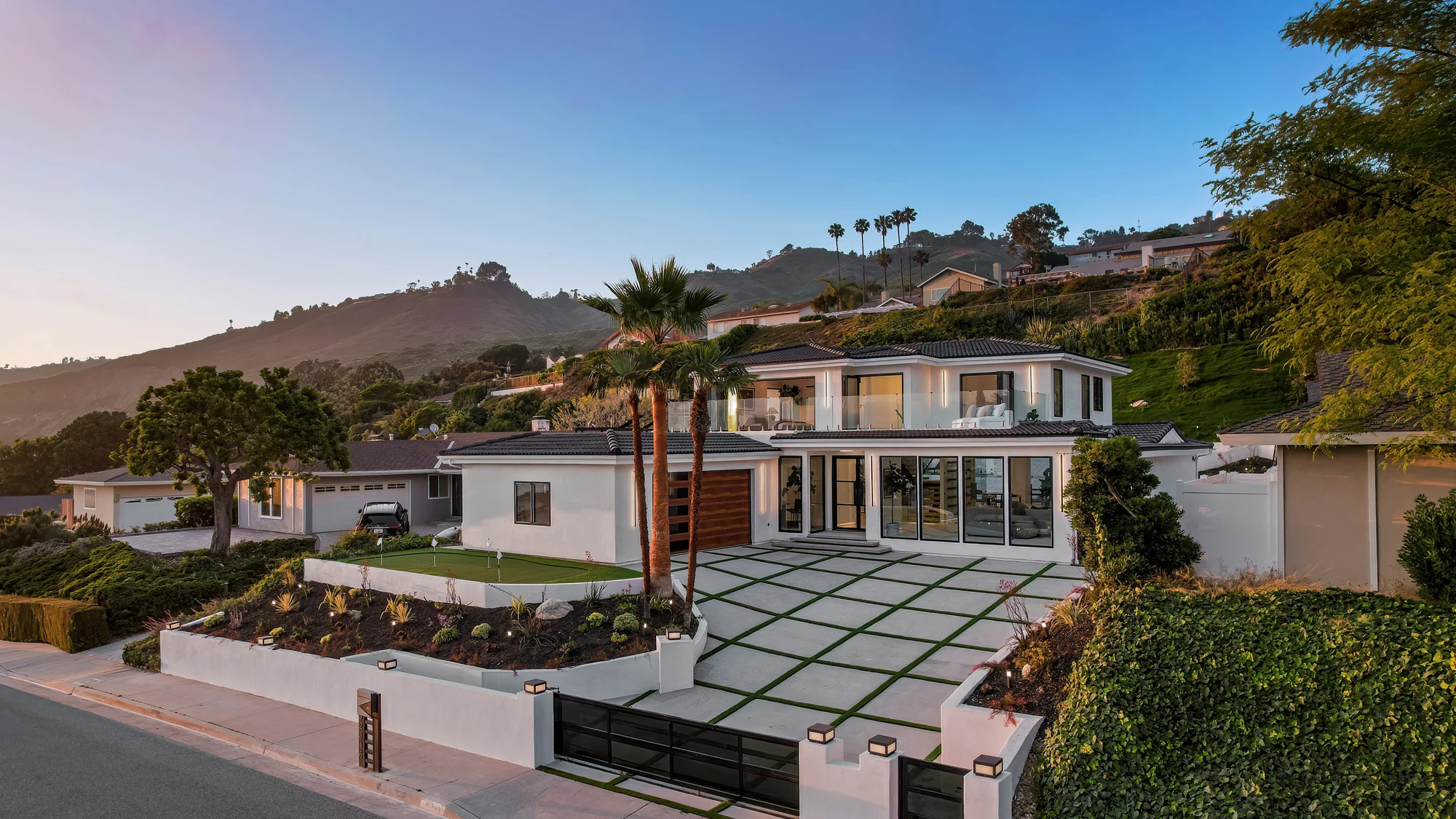 The finished white house at dusk on its Rancho Palos Verdes hillside, dark flat-tile roof, warm-lit glass across the ground floor and a paver-and-turf motorcourt, with the green ridge rising behind
