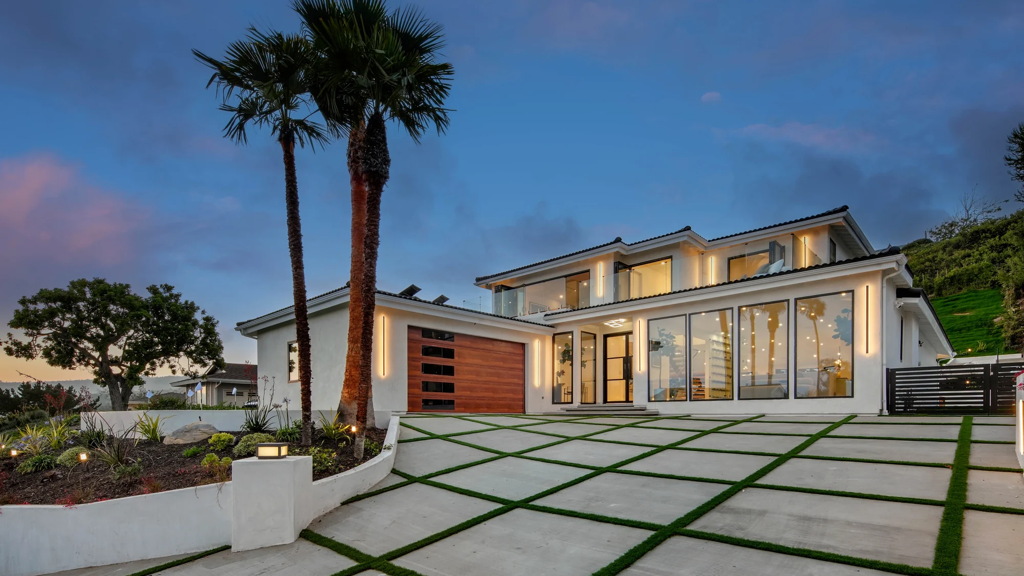 Front of the house at dusk with two mature fan palms, a low white wall, a black slatted driveway gate and a grid of pale pavers set into strips of green