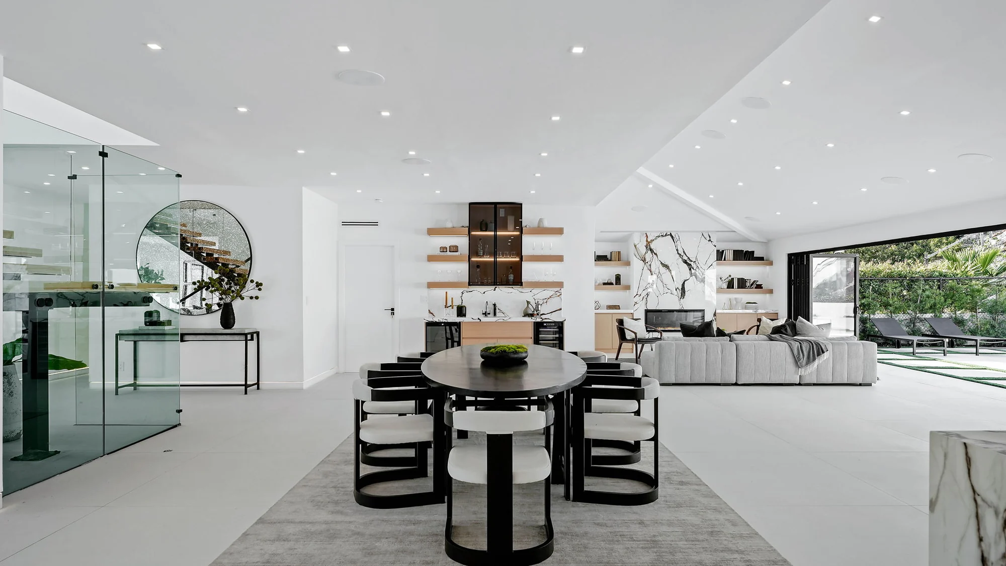 The open dining and living level in white, with a glass-walled wine room, floating oak shelving, a marble bar and the pocketing glass to the terrace standing open at the far end
