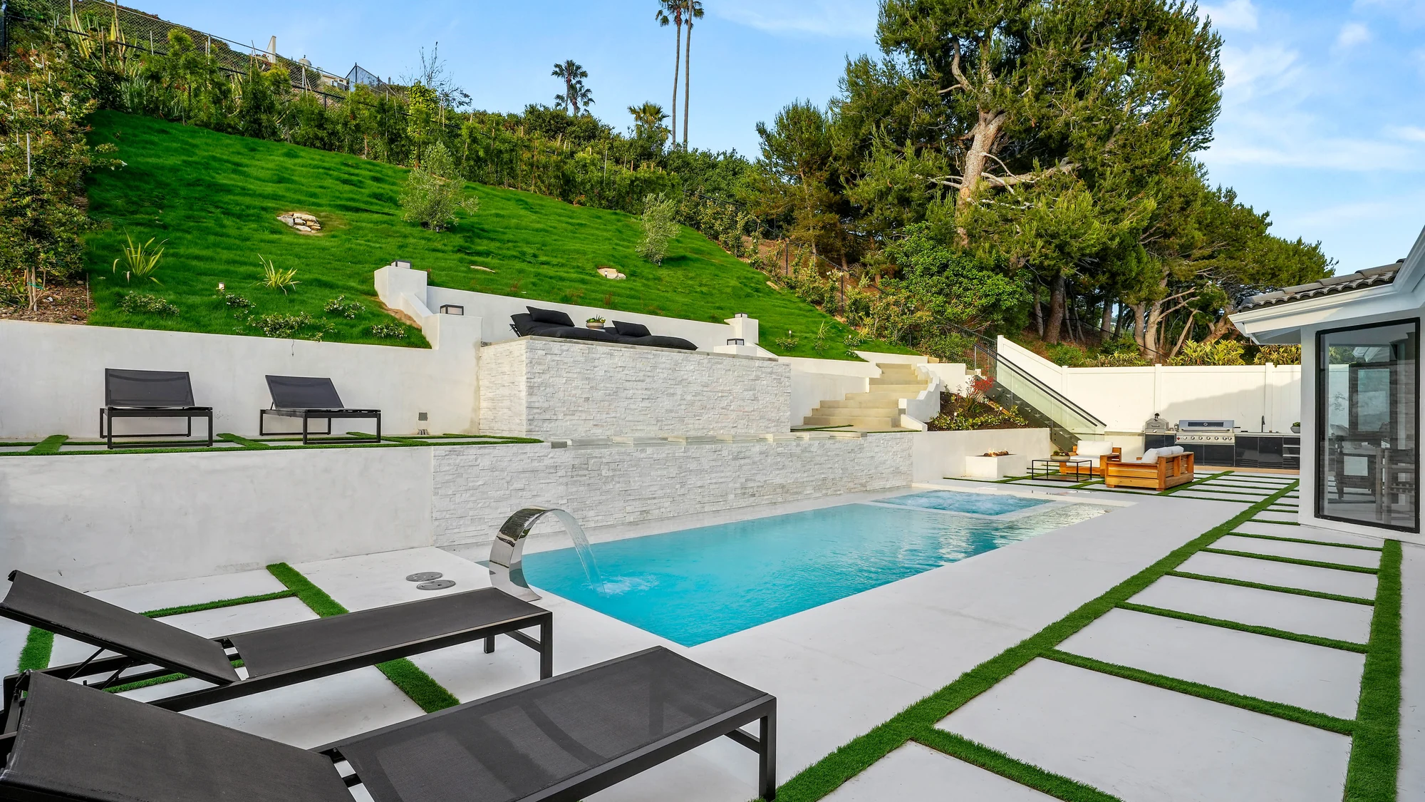 Rear garden with a rectangular pool and spa, a white stacked-stone wall with a sheer water spout, and a terraced green lawn climbing the hillside above