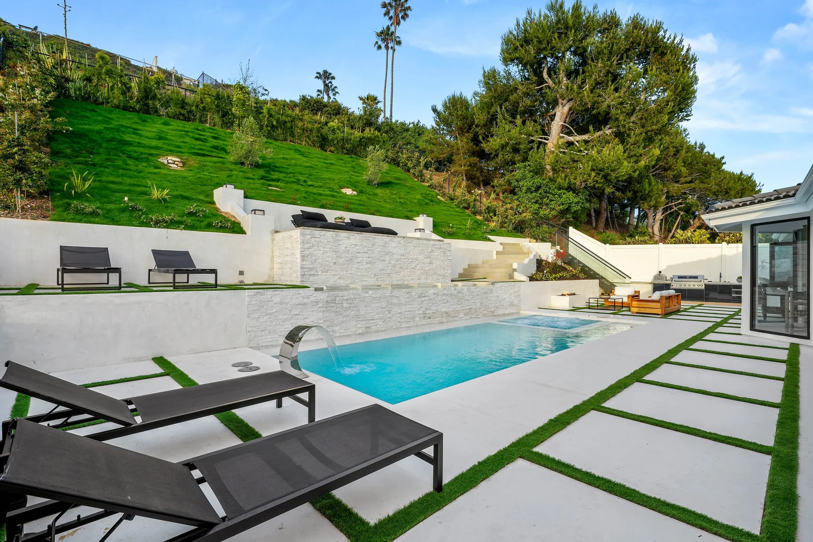 The rebuilt rear garden with a rectangular pool and spa, white board-formed and stacked-stone retaining walls, a sheer water spout, pavers set in turf, black loungers and a terraced green lawn rising up the hillside behind