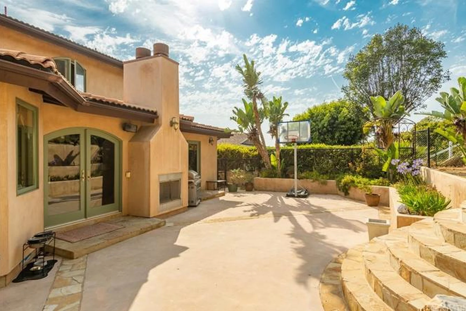 The original rear yard: a flat sand-coloured concrete slab, a stucco chimney mass with an outdoor fireplace, green arched French doors, a portable basketball hoop, banana palms and curved flagstone steps up to a raised planter